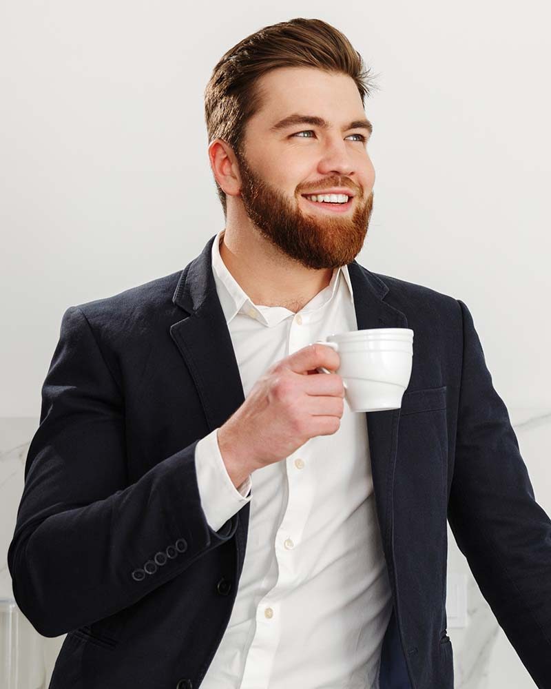Portrait of a smiling young businessman dressed in suit standing and drinking coffee indoors