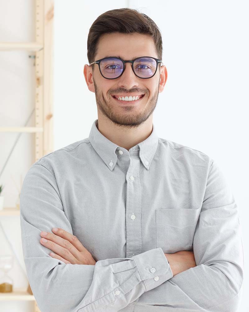 Portrait of young business man wearing gray shirt and glasses, stand with crossed arms in office, looking confident, professional, smiling positively at camera