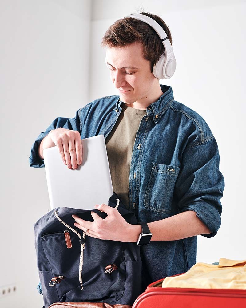 Cheerful young student in wireless headphones packing tablet in satchel while going to travel