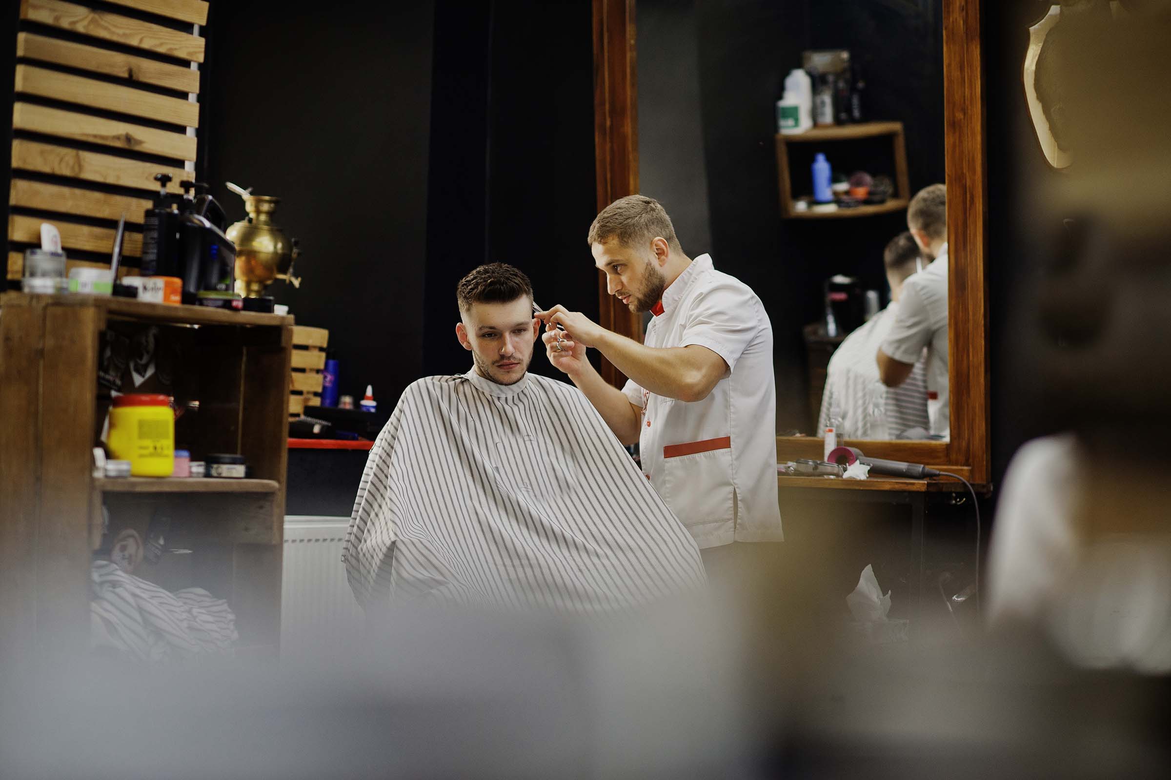 Young bearded man getting haircut by hairdresser while sitting in chair at barbershop. Barber soul.