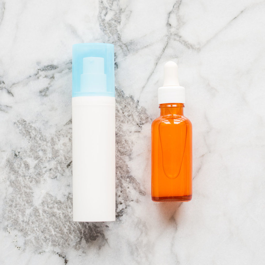 Face cream in white pump dispenser and skin serum in orange glass bottle on marble board. Skin care products concept